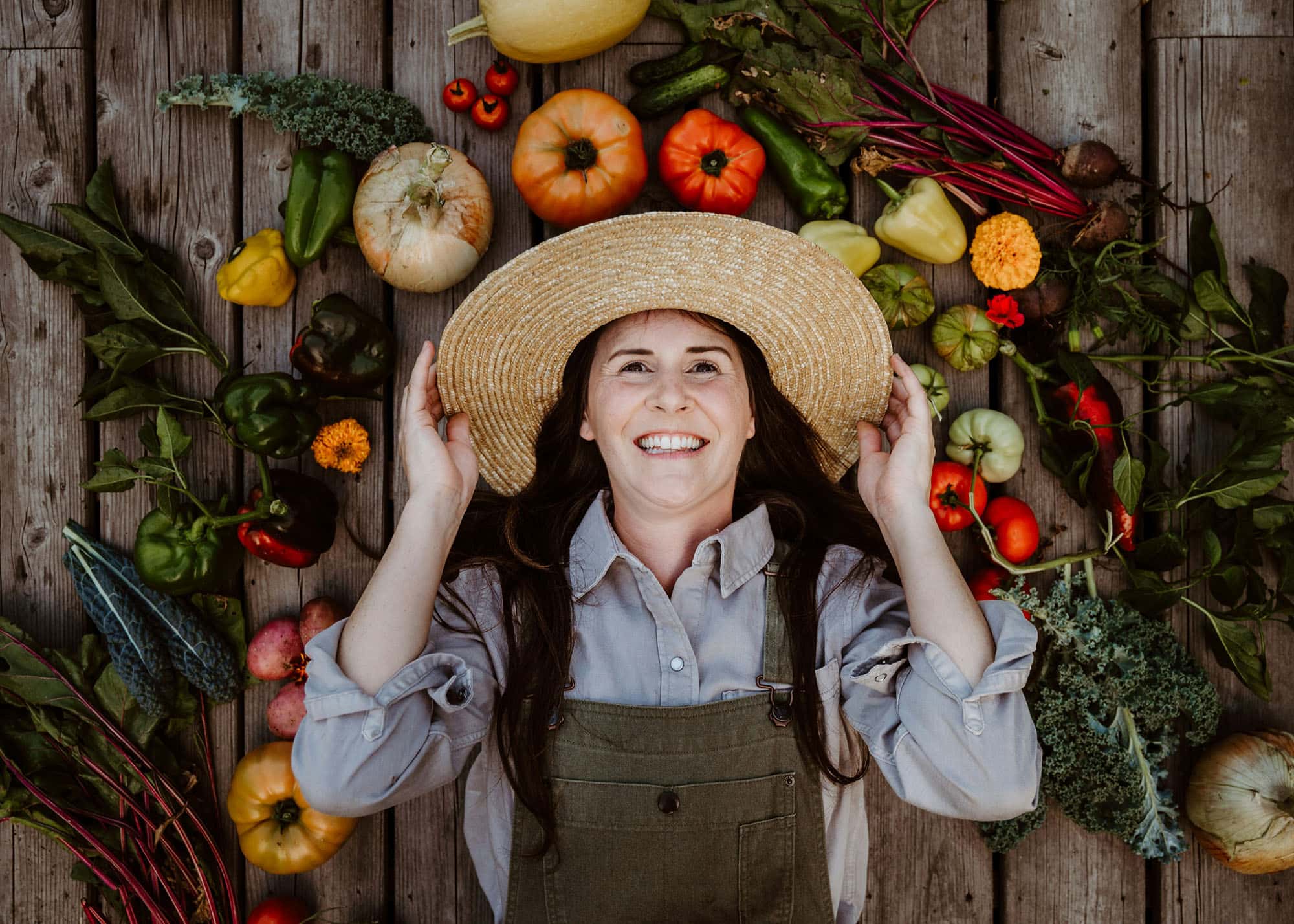 A laughing farmer in a straw hat lying on wooden boards, surrounded by tomatoes, peppers and other fresh vegetables.