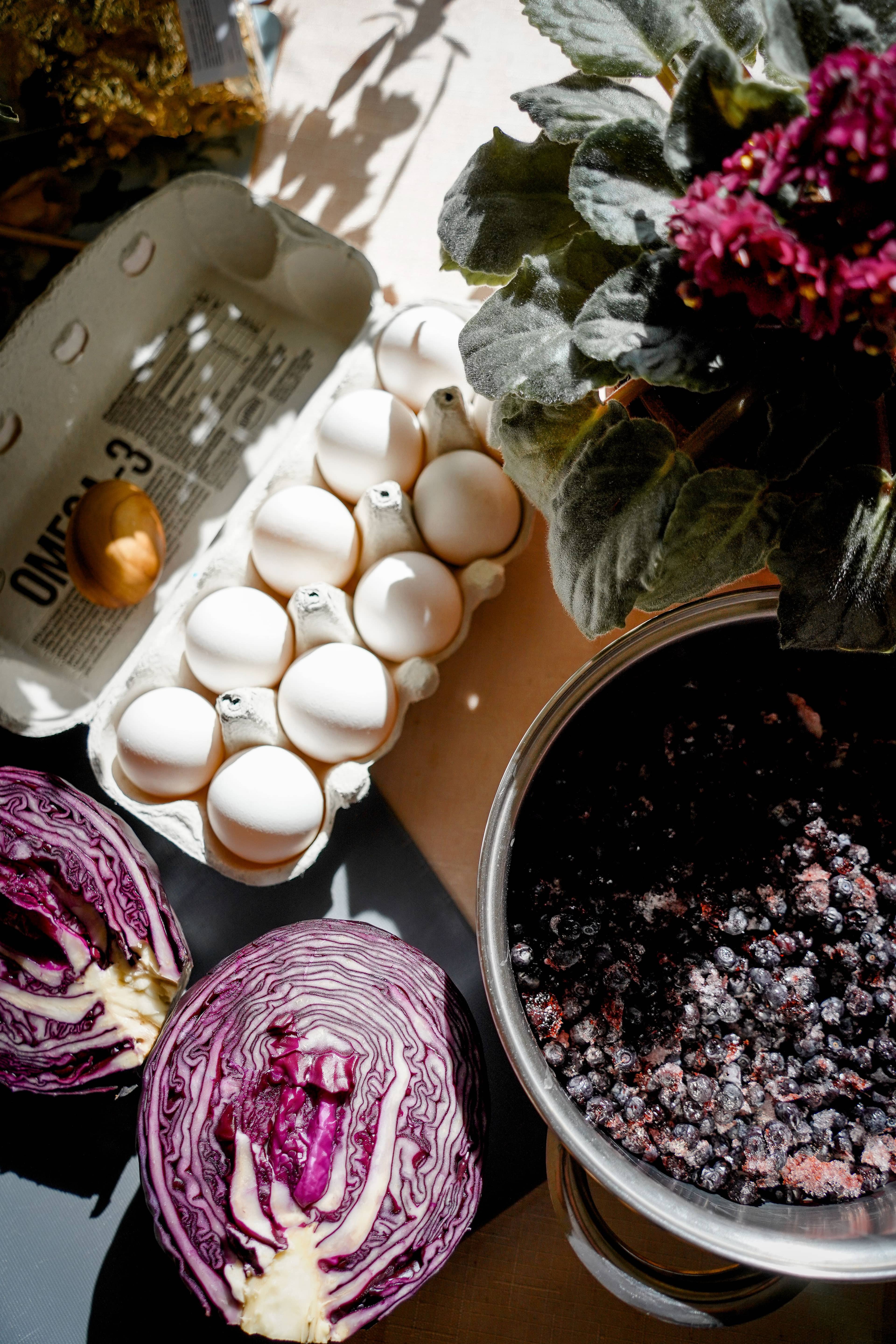 A sunlit tabletop with a carton of eggs, halved red cabbage, and a bowl of blueberries.